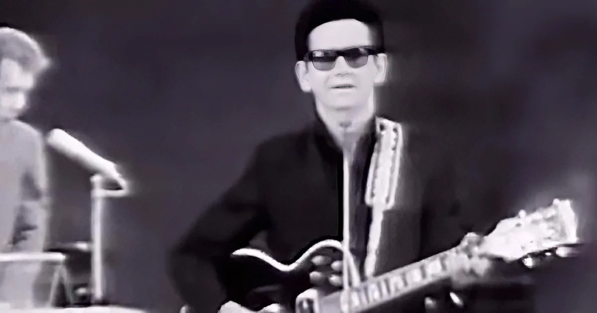 A man in sunglasses plays a guitar on stage, wearing a dark suit, with another person in the background.