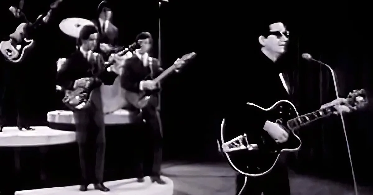 A man in sunglasses plays guitar on stage with a band, black-and-white image.