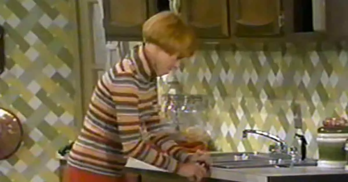 A child in a striped shirt stands at a kitchen sink, washing dishes.
