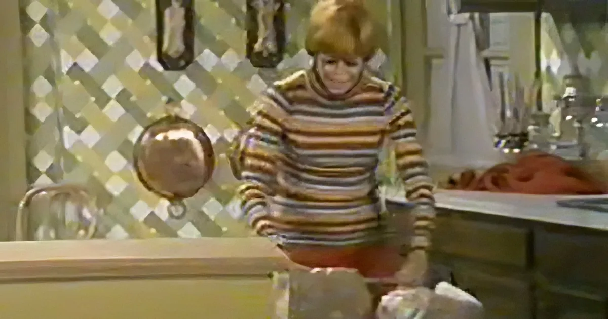A person in a striped shirt stands in a kitchen, holding a pot, with a checkered wall and utensils in the background.