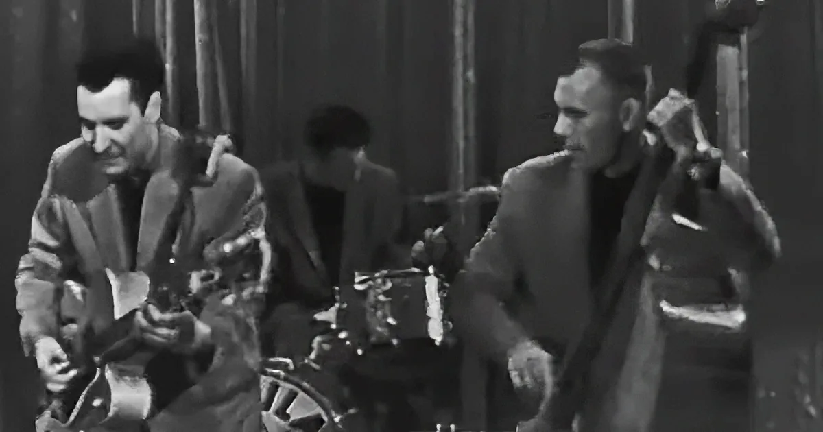A man in a suit plays guitar while another in a suit sits nearby, both in a dimly lit room.