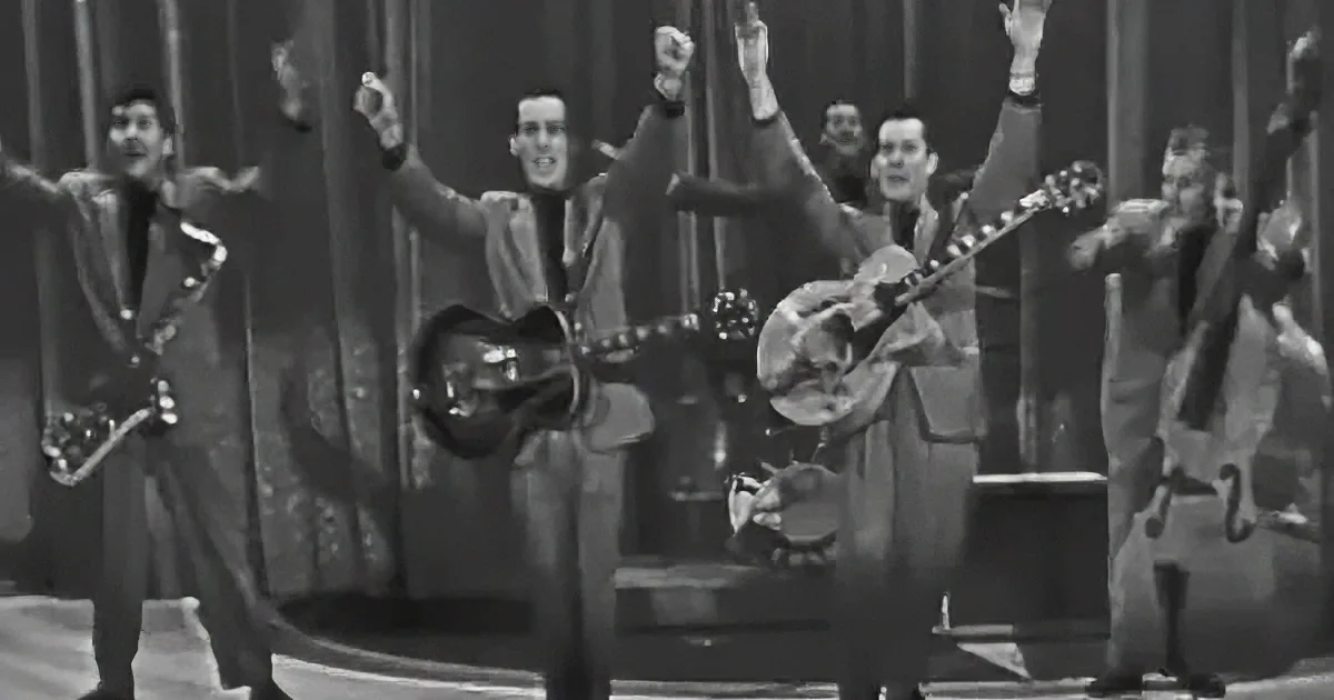 A musician in a suit holds a guitar, surrounded by a band in a lively performance setting.