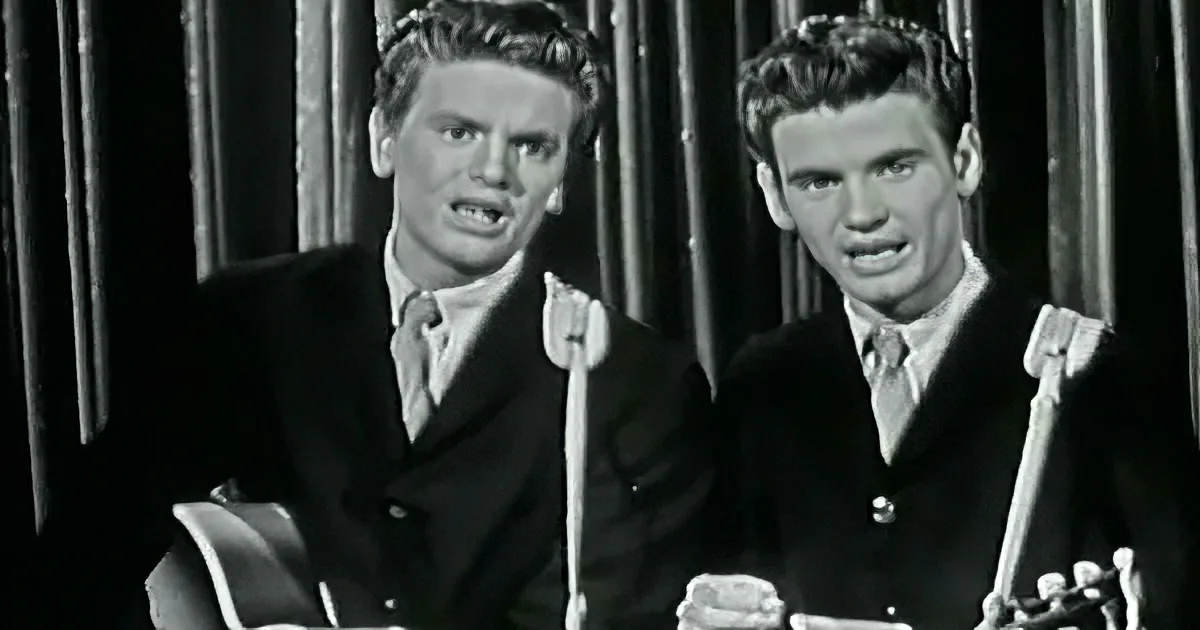 Two men in suits with guitars, one singing, black-and-white image.