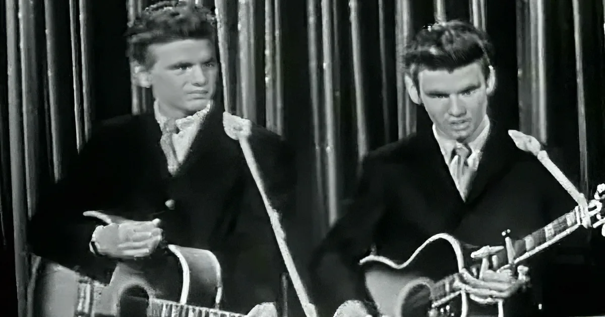 Two men in suits play guitars, one holding a microphone, against a curtain backdrop.
