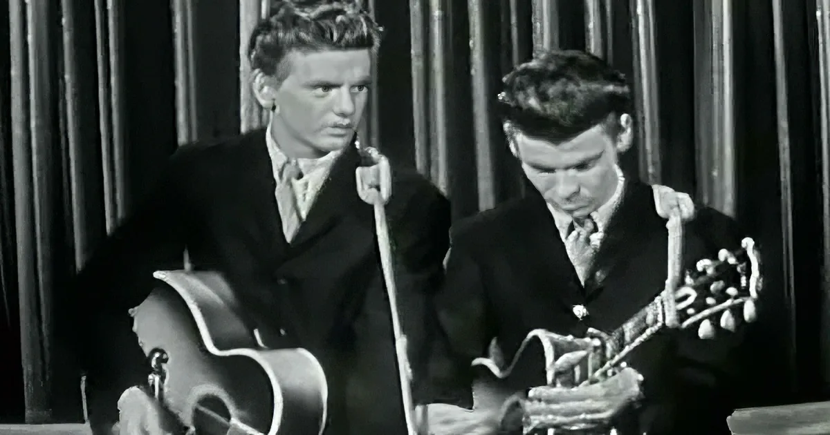 Two men in suits play guitars on stage, one with a microphone.