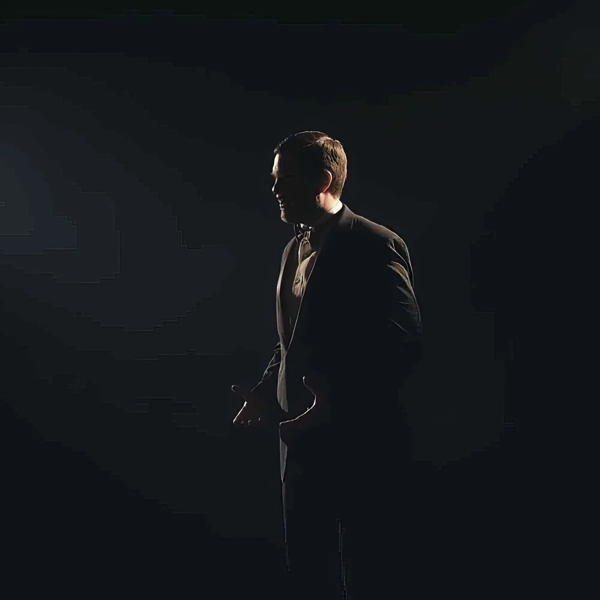 A man in a suit and bow tie stands in silhouette against a dark background, holding a cane.