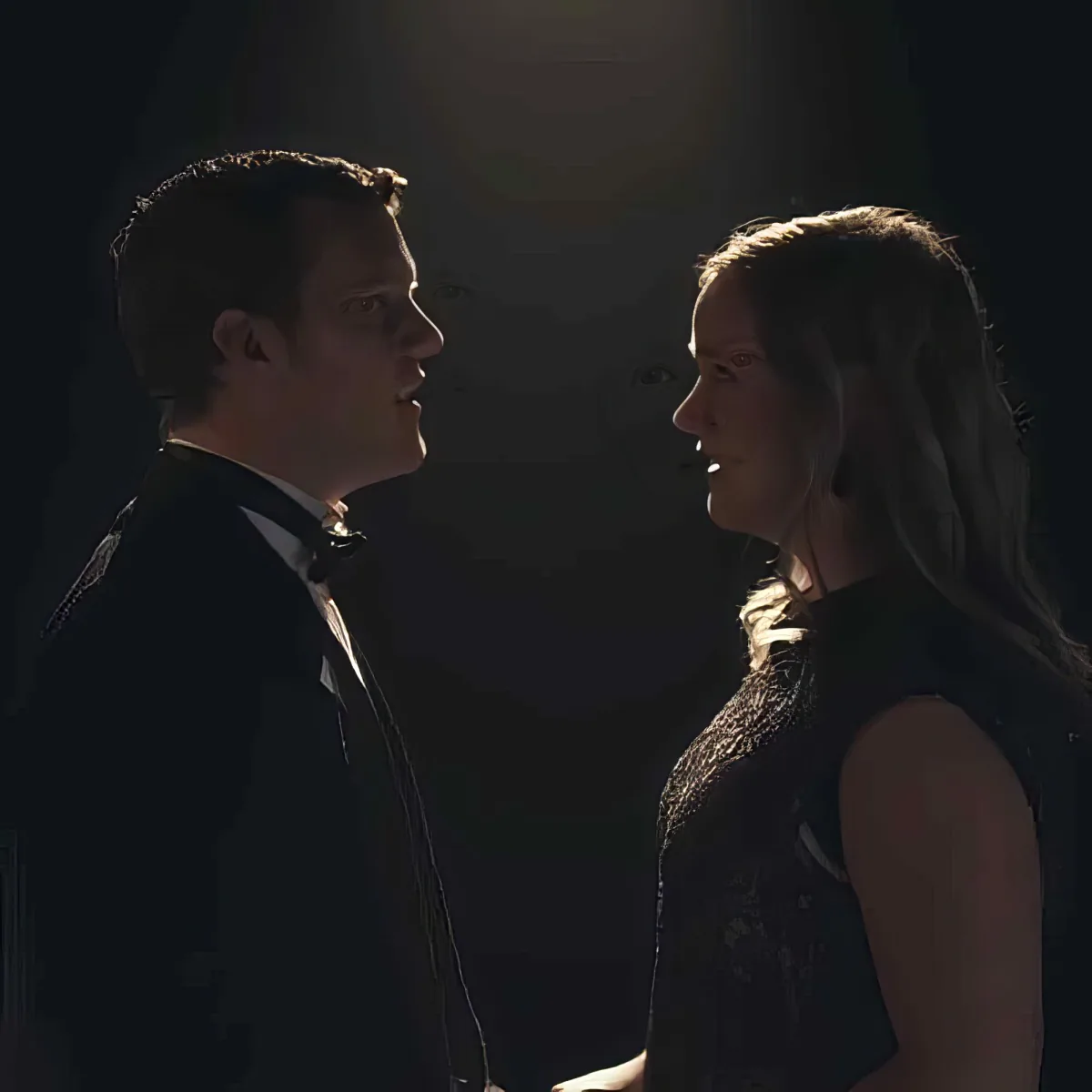 A man in a suit and a woman in a black dress are facing each other in a dimly lit room.