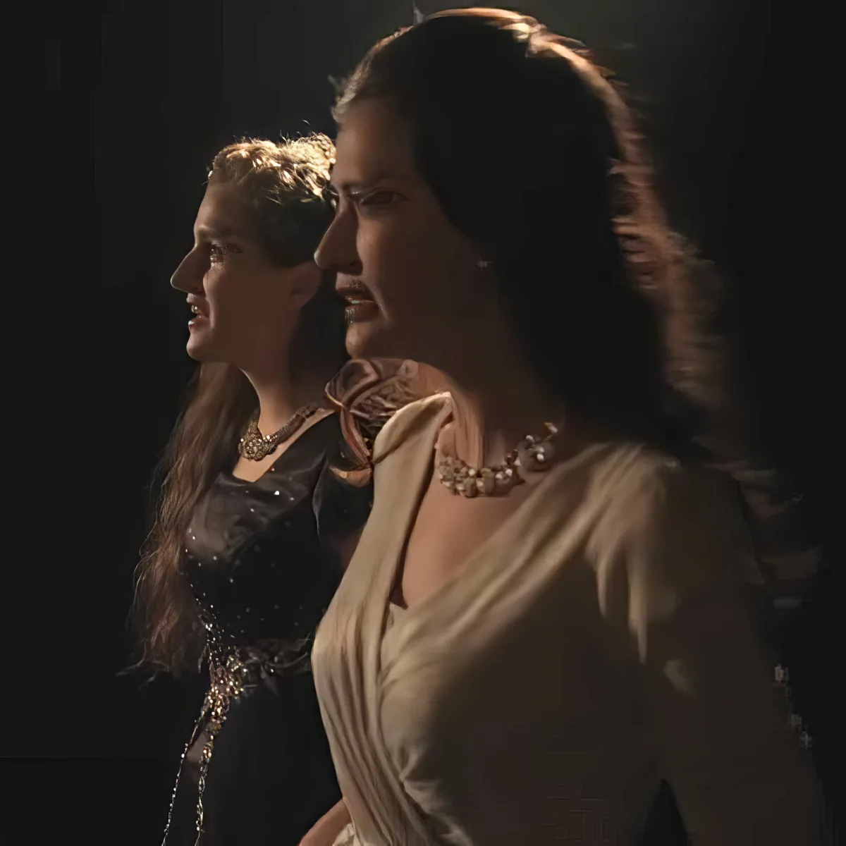 A woman in a white robe stands beside another woman in a black dress, both with long hair, in a dimly lit setting.