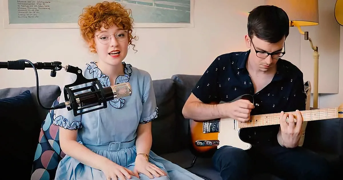 A woman in a blue dress sings into a microphone while a man plays a guitar on a couch.