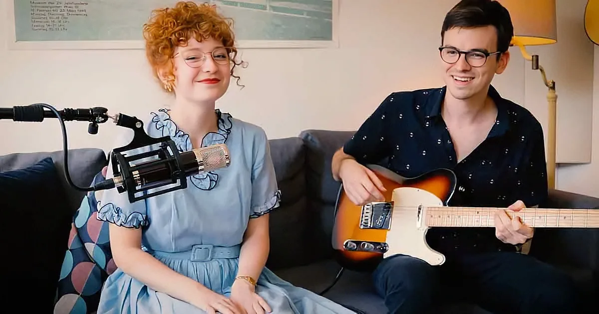 A woman in a blue dress sits next to a man playing a guitar, both smiling, with a microphone in front of her.