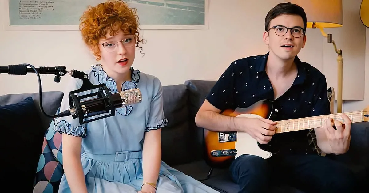 A woman sings into a microphone while a man plays guitar on a couch.