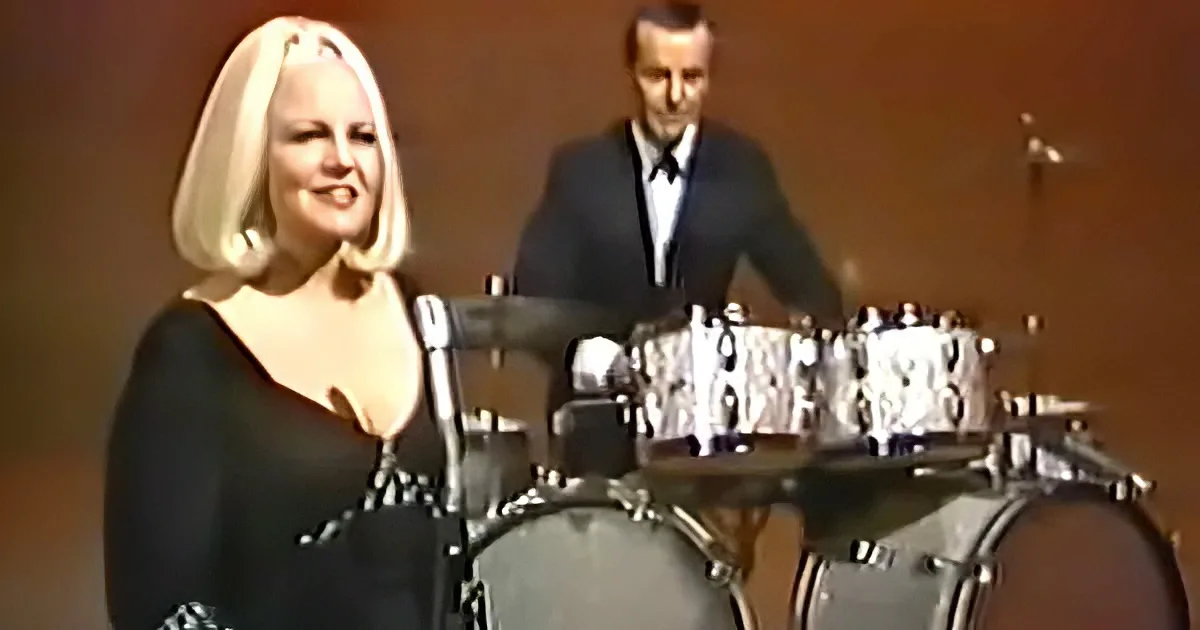 A woman with blonde hair and a black dress smiles while playing drums, with a man in a suit behind her.