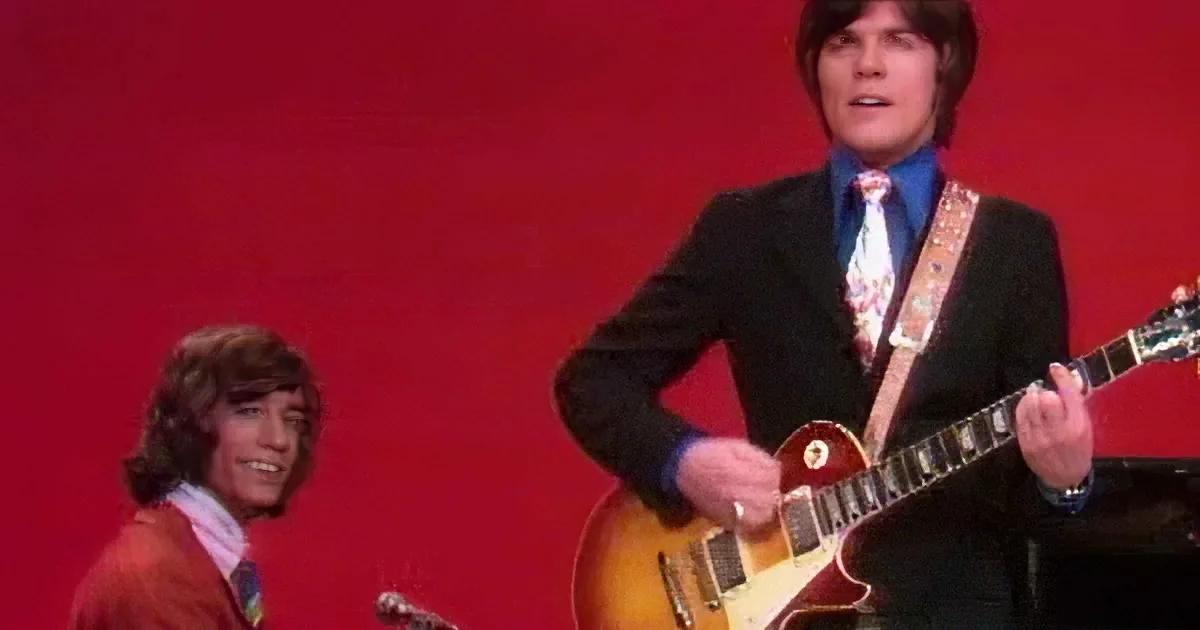 A man in a suit plays a red guitar on stage, with another person in the background.