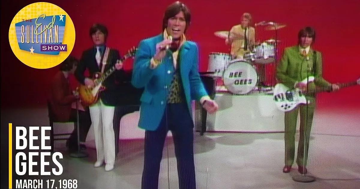 A person with shoulder-length dark hair, wearing a blue and yellow outfit, smiles against a red background.