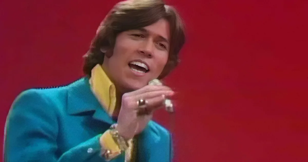 A person in a teal suit with a yellow collar sings into a microphone against a red background.