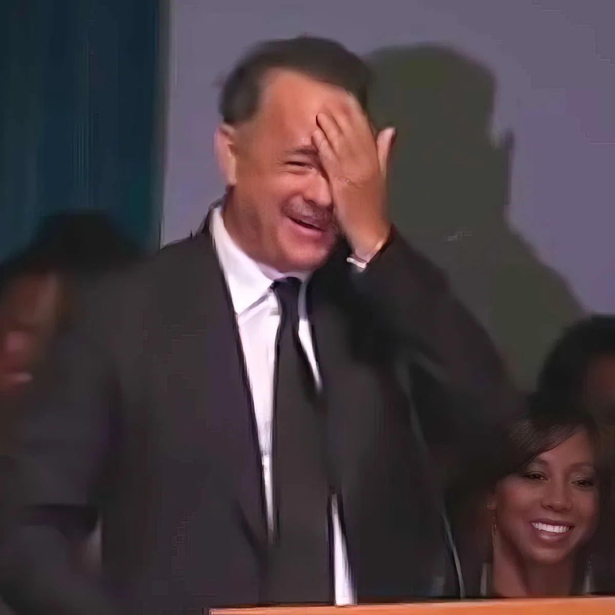 A man in a suit covers his face with his hand, smiling, with blurred people in the background.