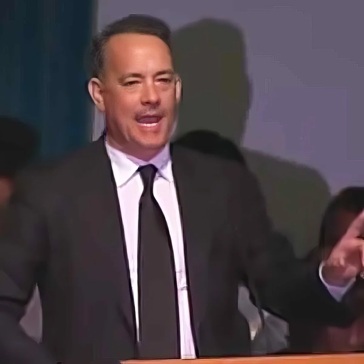 A man in a suit and tie is smiling while standing at a podium, with blurred figures in the background.