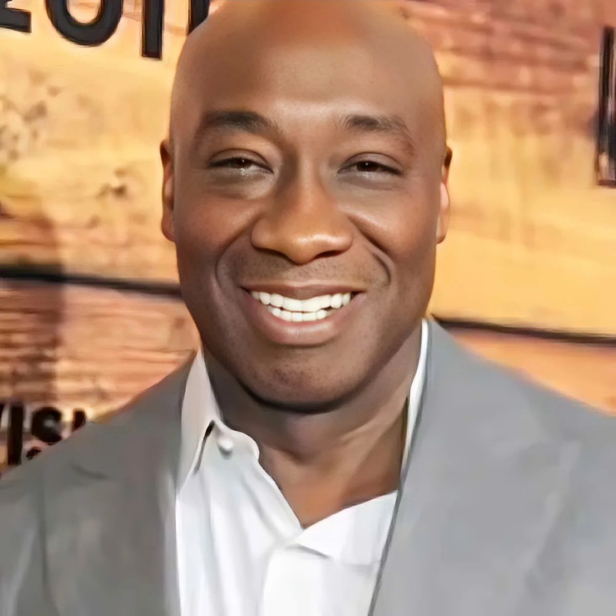 A smiling man in a light gray suit jacket and white shirt, with a brick wall background.