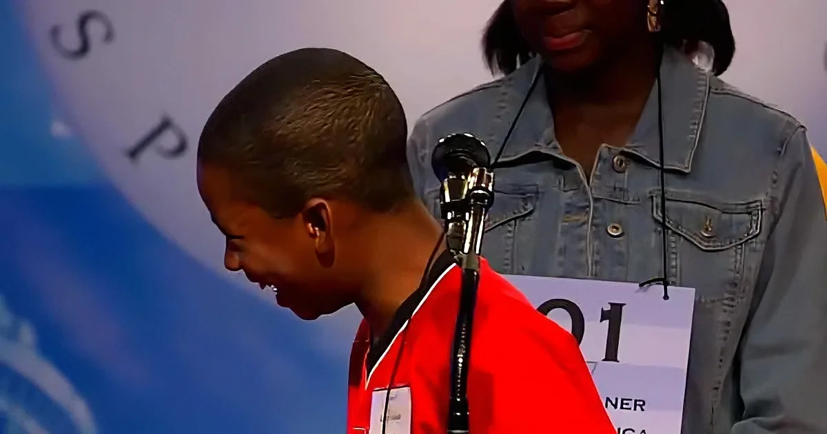 A young boy in a red jacket stands at a microphone, smiling, with a woman in a denim jacket behind him.