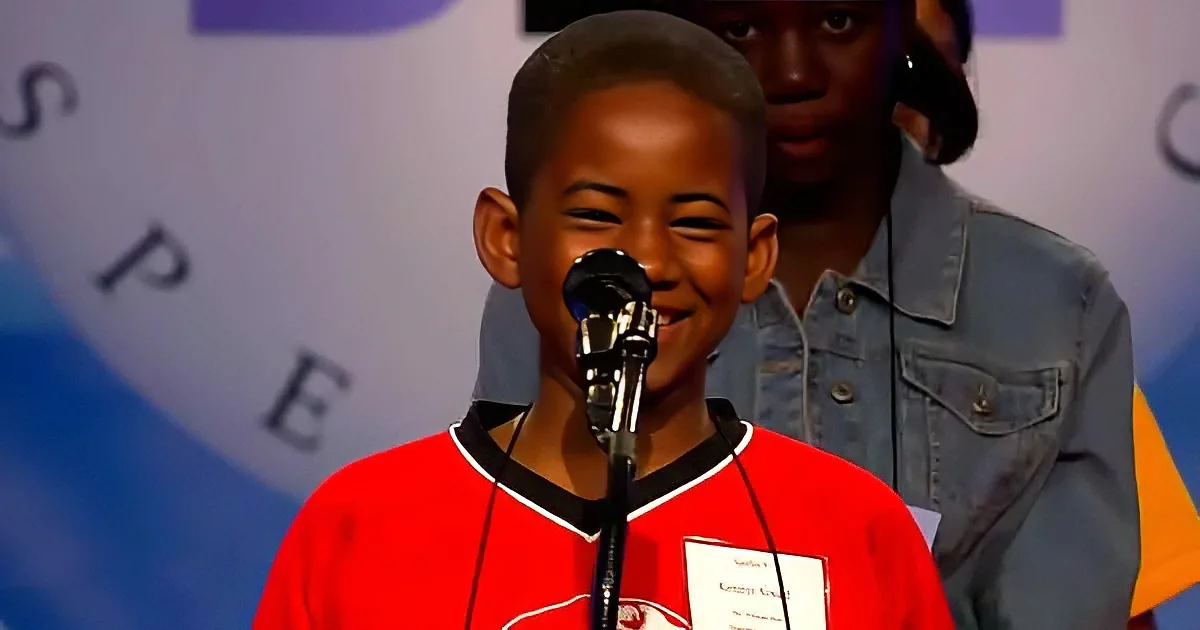 A young boy in a red shirt smiles while speaking into a microphone at a podium.