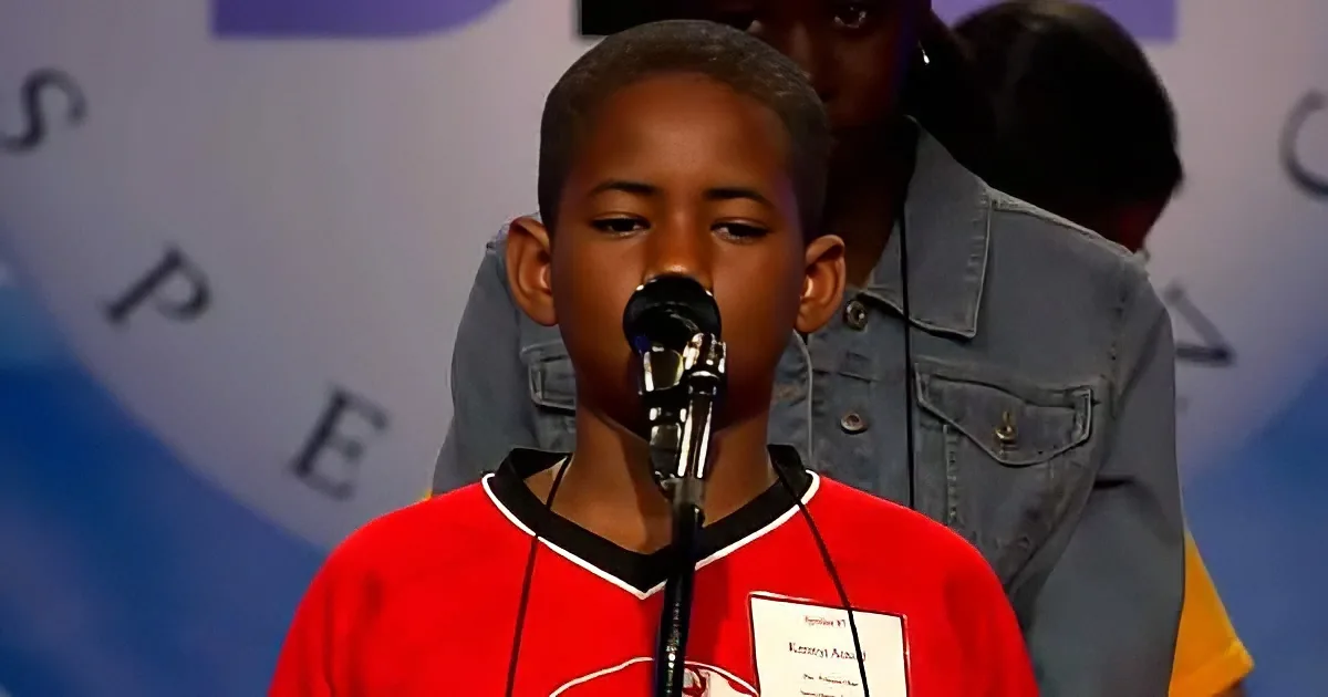 A young boy in a red shirt stands at a microphone, wearing a name tag, with a blurred background.