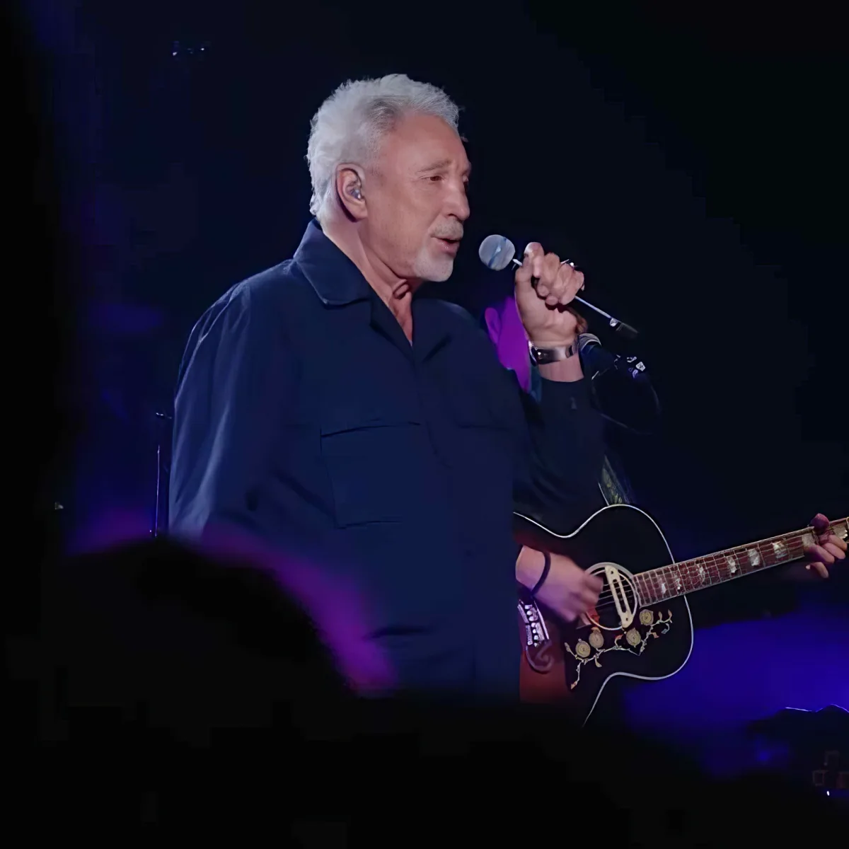 A man with white hair and a beard sings into a microphone while holding an acoustic guitar on stage.