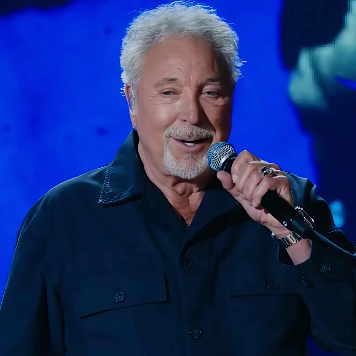 An older man with white hair and a mustache holds a microphone, wearing a dark jacket, against a blue background.