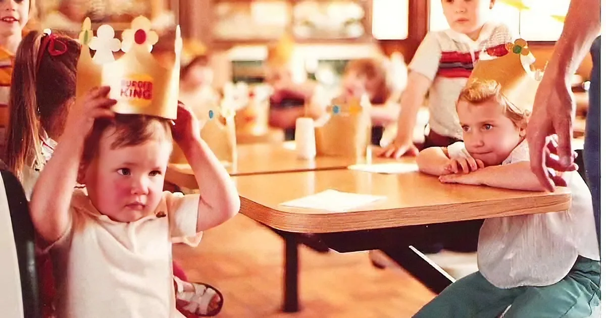 A baby adjusts a Burger King crown while sitting at a table in a restaurant with other children.