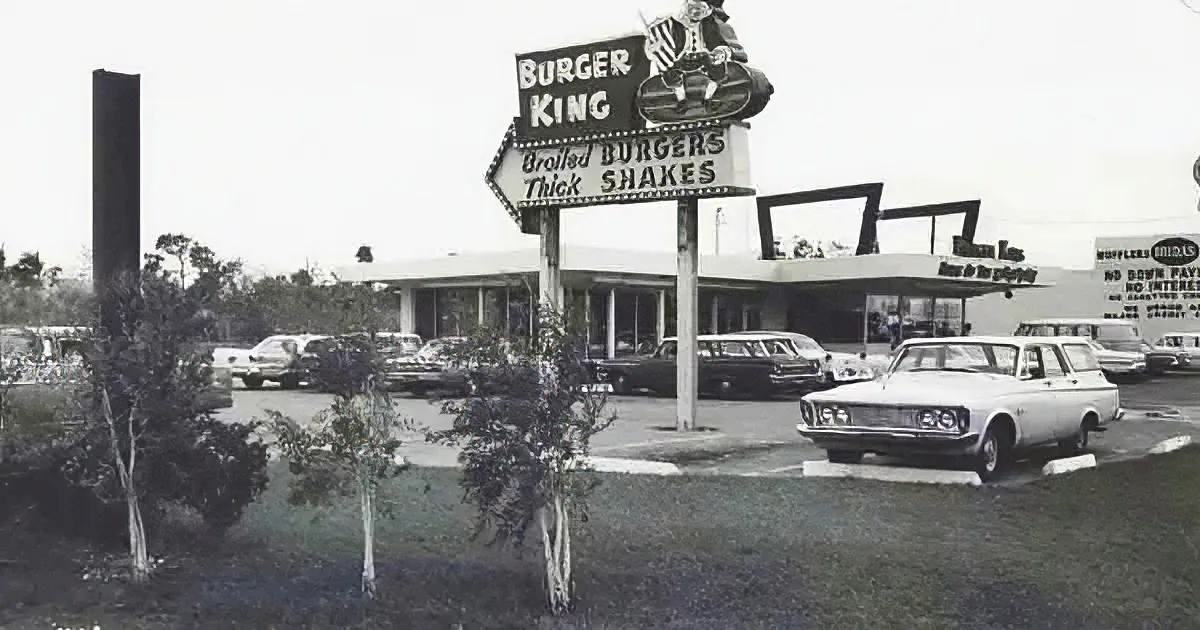 A vintage Burger King sign advertises broiled burgers and shakes, with cars parked in front of the restaurant.
