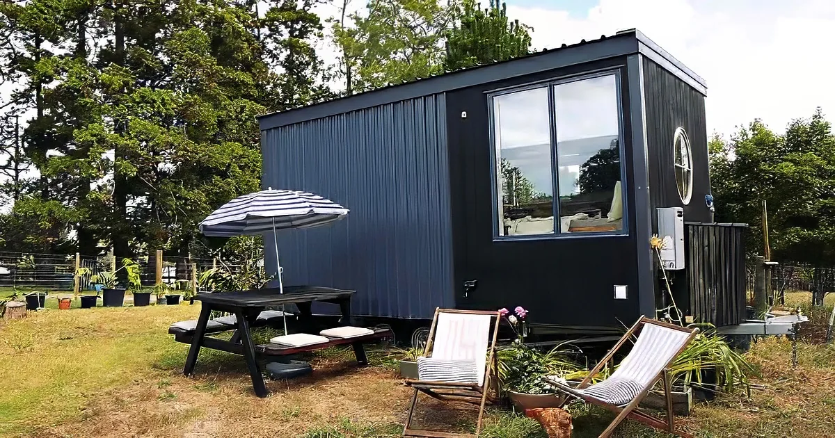 A black shipping container house with large windows, a picnic table, and two lounge chairs in a grassy yard.