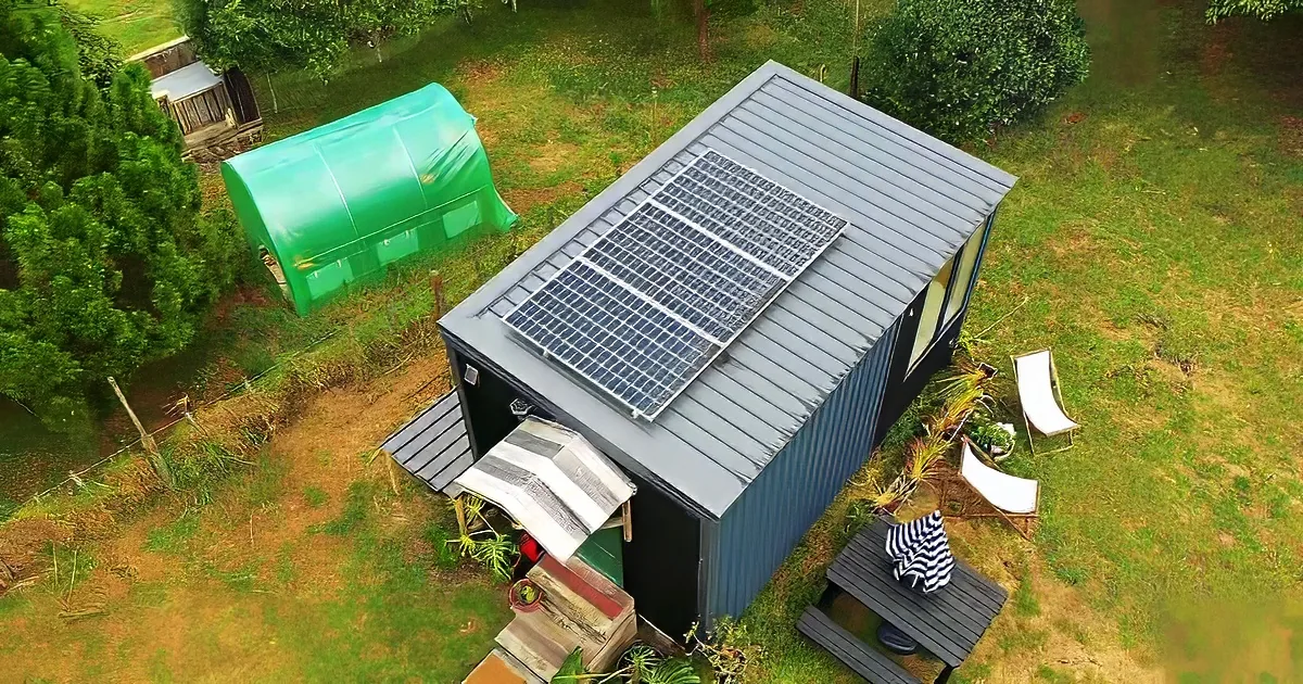 A small house with a solar panel on its roof sits in a grassy yard with a green shed and picnic table nearby.