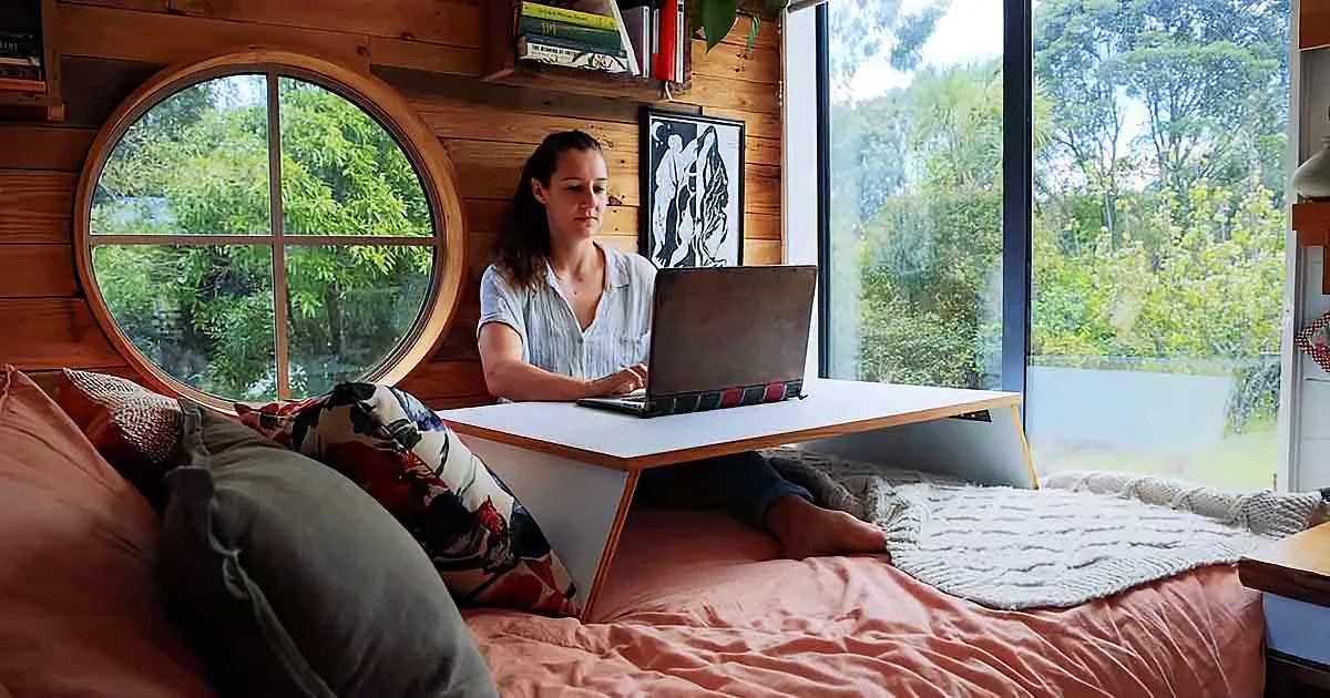A woman works on a laptop on a bed in a cozy, wooden cabin with a large window.