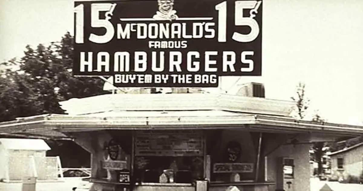 A vintage McDonald's sign advertises 15 famous hamburgers, "Buy 'em by the bag," above a classic drive-thru window.