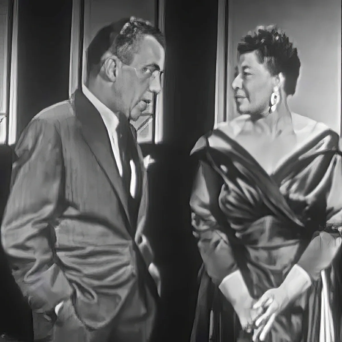 A man in a suit and a woman in a dress are engaged in conversation, with the man appearing to be listening intently.