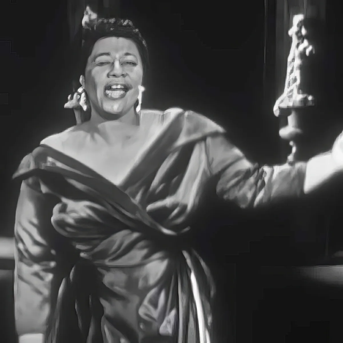 A woman in a flowing robe, holding a staff, and wearing large earrings, appears to be singing or speaking passionately.