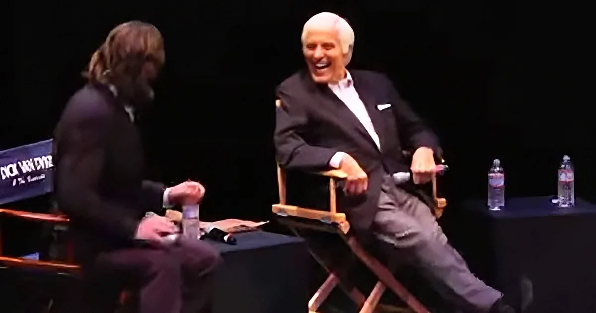An older man in a suit sits in a director's chair, laughing, while another person in a suit sits beside him.