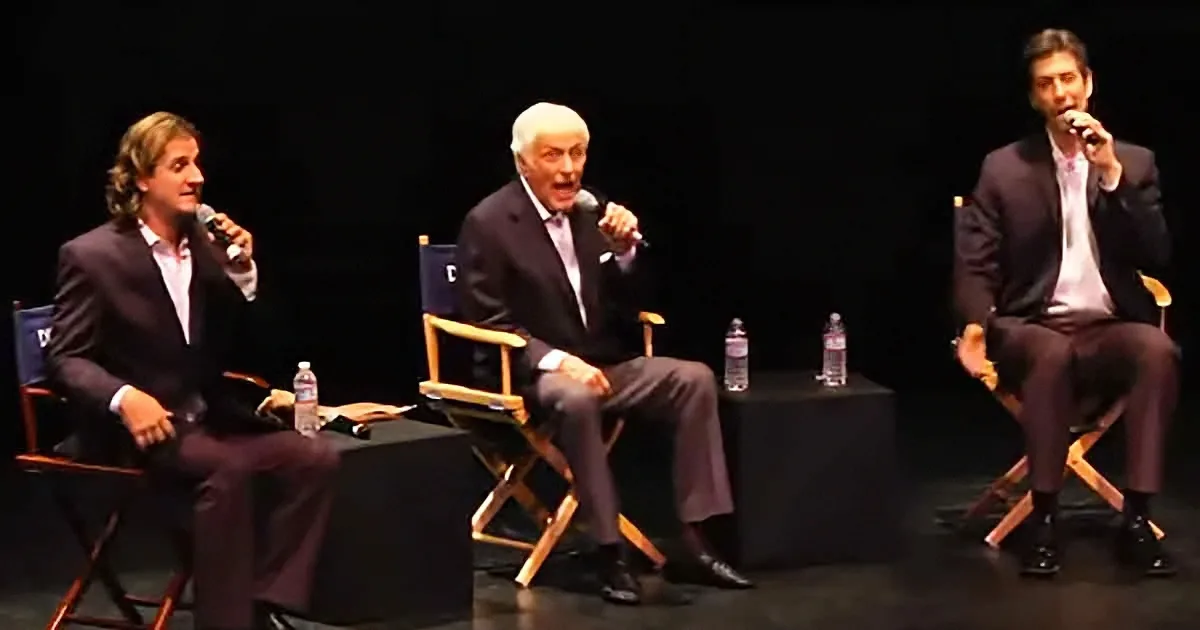 Three men in suits sit on stage, each holding a microphone, engaged in a discussion.
