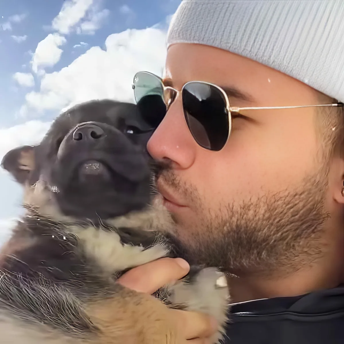 A man wearing a beanie and sunglasses holds a fluffy puppy against a blue sky with clouds.