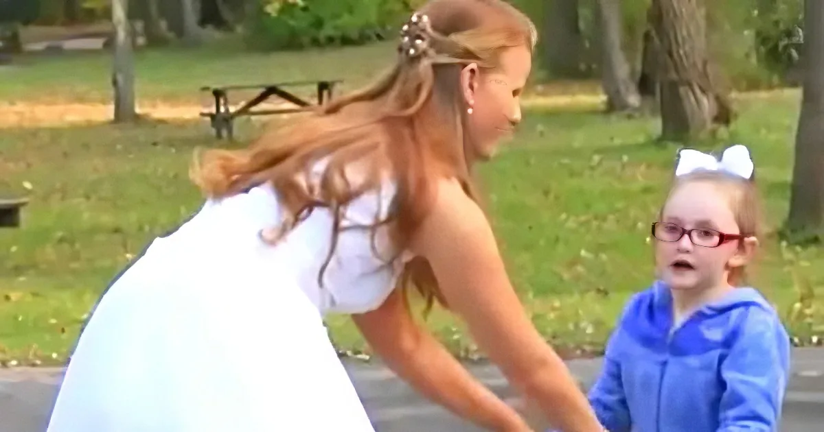 A woman in a white dress bends forward, while a young girl in a blue hoodie looks on in a park setting.