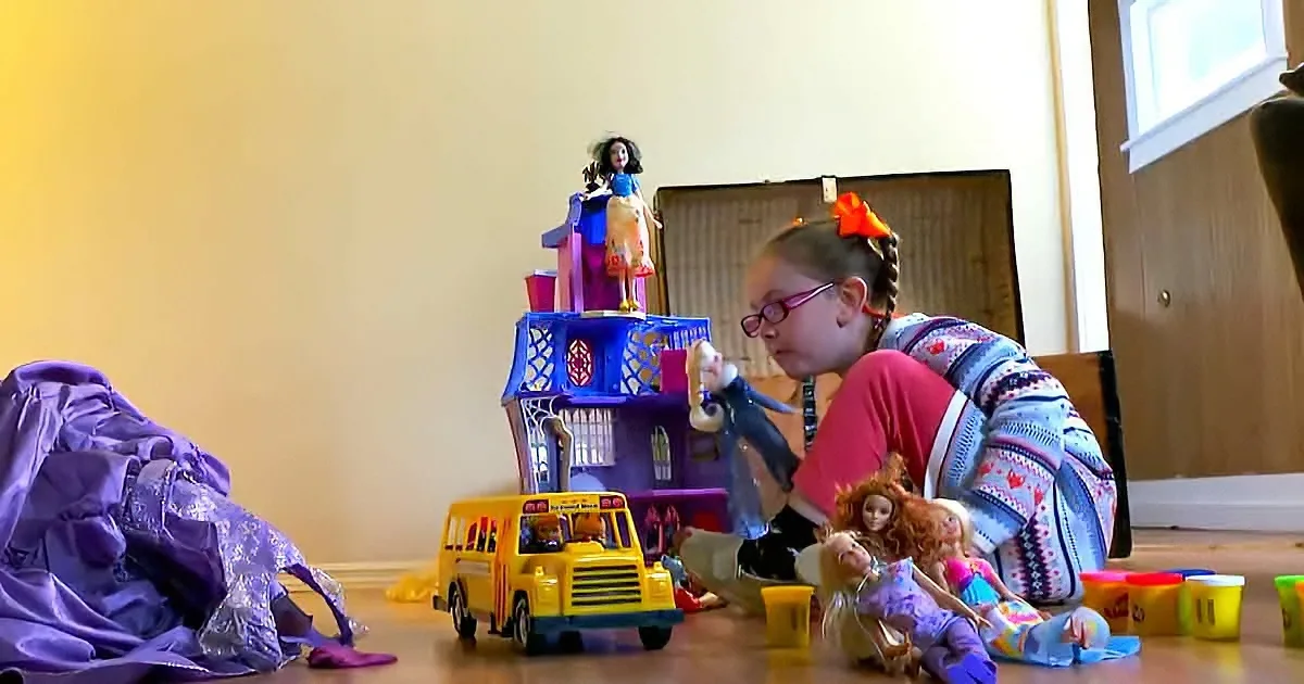 A young girl plays with dolls and a school bus on the floor, surrounded by toys and clothes.