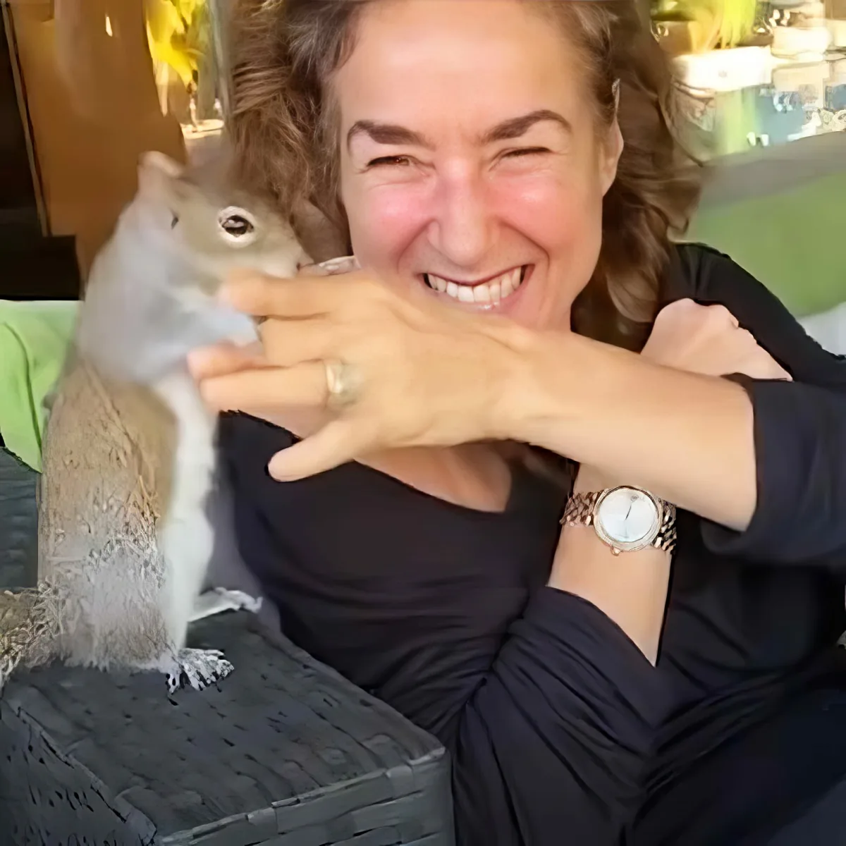 A woman is smiling widely while holding a squirrel on her lap, both appearing happy.