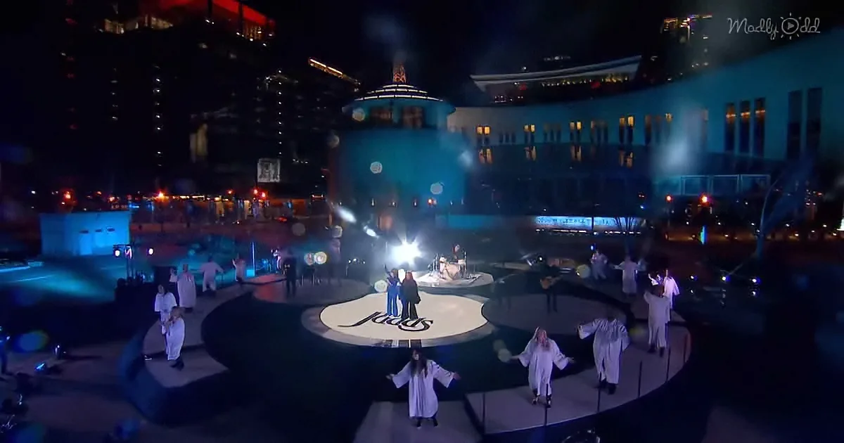 A group of performers in white robes stand on a circular stage with a "Kiss" logo, illuminated by bright lights.