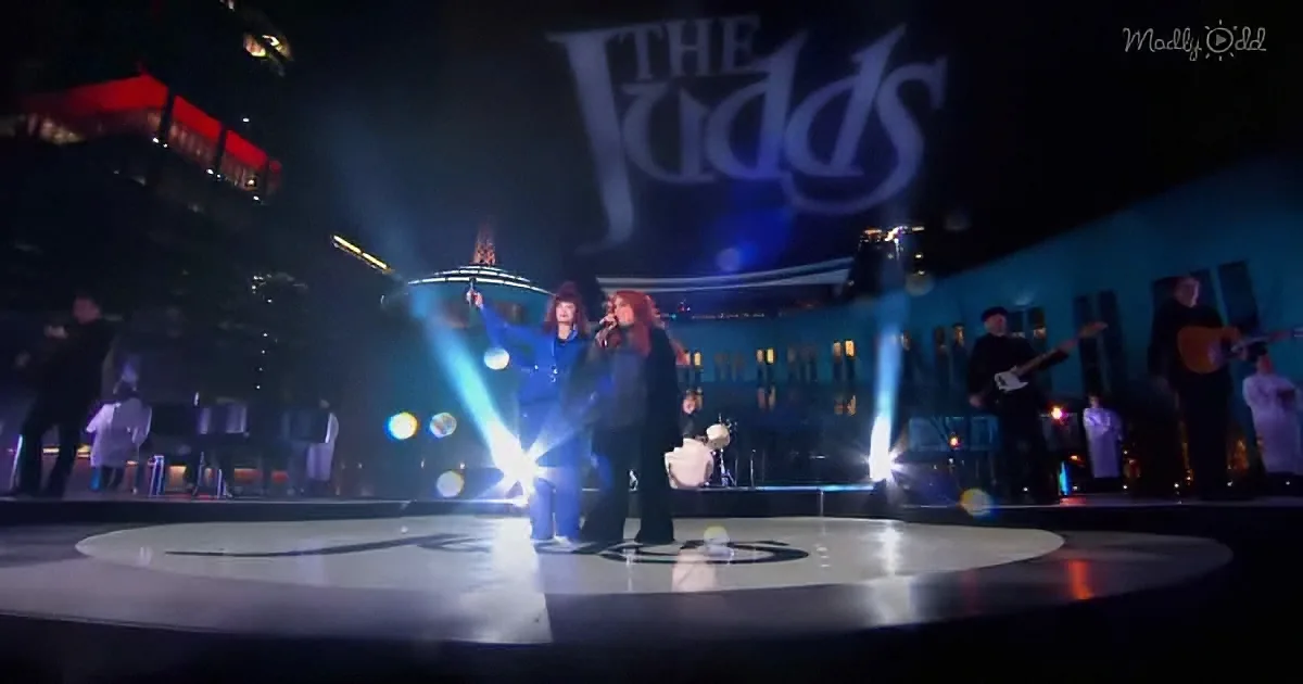 Two women sing on stage with "The Vudds" logo, surrounded by musicians and bright stage lights.