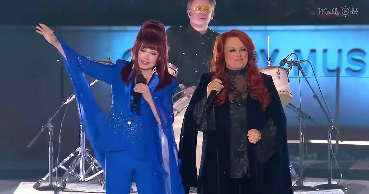 Two women in vibrant outfits perform on stage, one in blue, the other in black, with microphones and a drummer in the...