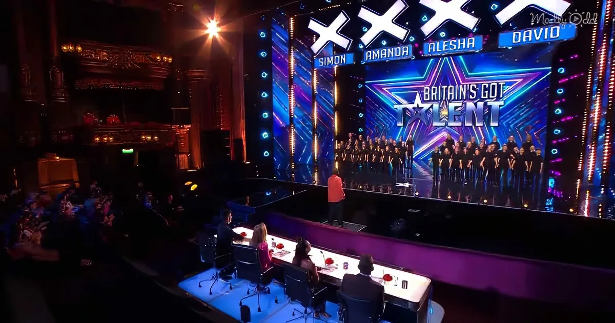 A choir performs on a brightly lit stage with judges seated at a table in the foreground.