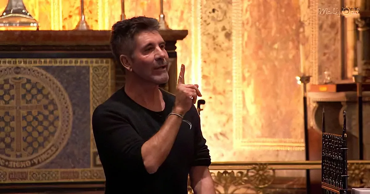 A man in a black shirt gestures with his right hand, standing in front of ornate, golden decor.