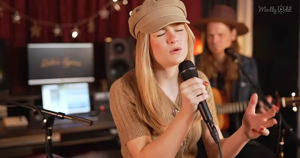 A woman in a hat sings into a microphone, holding a guitar, with a recording setup in the background.