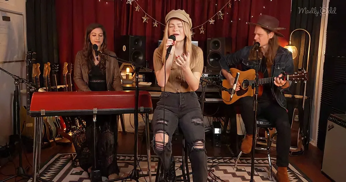 A woman sings into a microphone while playing a keyboard, accompanied by a guitarist and another woman in a studio setting.