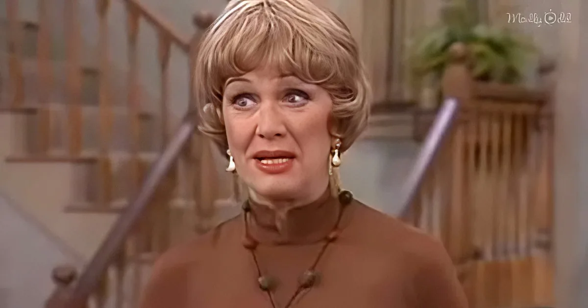 A woman with blonde hair and earrings, wearing a brown top, stands indoors near a staircase.