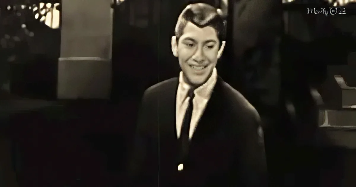 A man in a suit and tie smiles, wearing a distinctive hairstyle, in a dimly lit setting.
